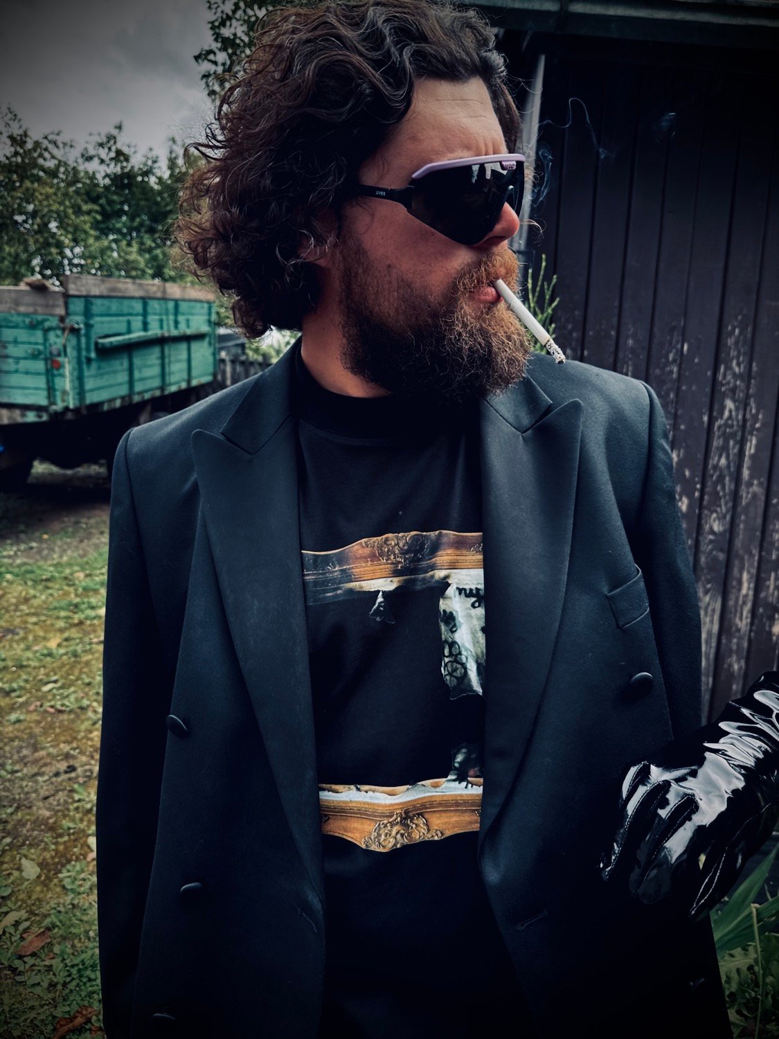 Barzdotas vyras su tamsiais akiniais rūko cigaretę lauke.