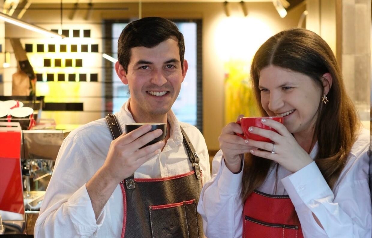 Vyras ir moteris su „Koffee Lab“ prijuostėmis šypsosi ir geria kavą iš puodelių.