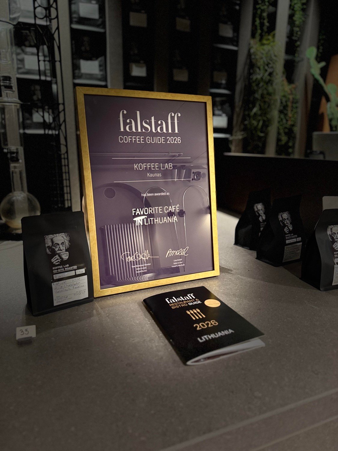 Rėmelis su „Falstaff Coffee Guide 2026“ apdovanojimu ir kavos maišeliai ant stalviršio.