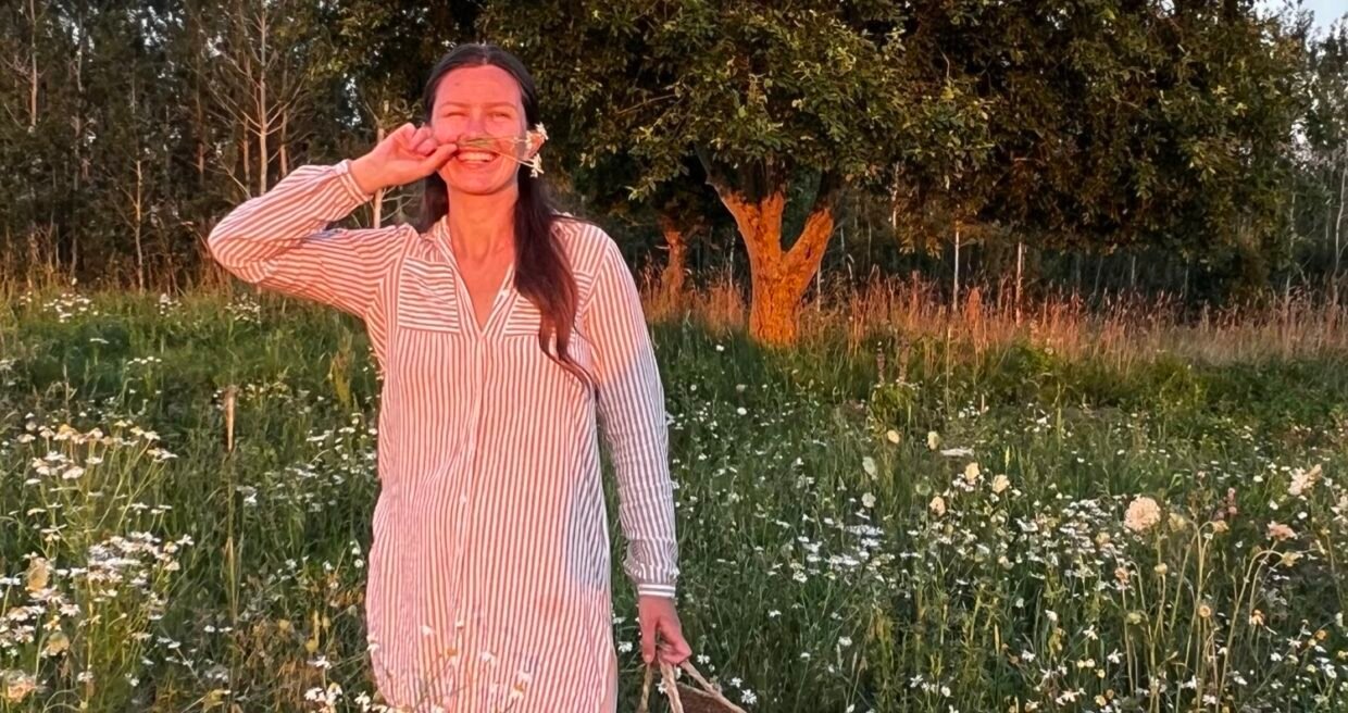 Gintarė Petrauskaitė besišypsanti gėlių pievoje su pinučiu krepšiu rankoje.