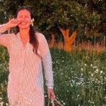 Gintarė Petrauskaitė besišypsanti gėlių pievoje su pinučiu krepšiu rankoje.