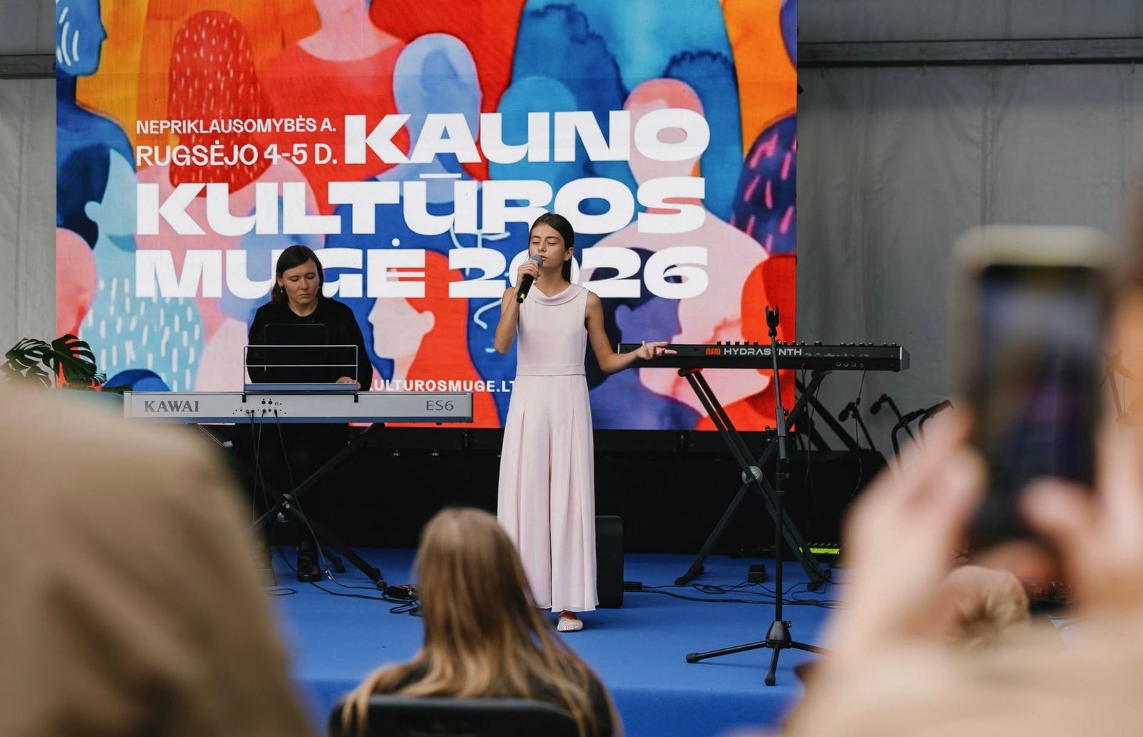 Mergaitė dainuoja Kauno kultūros mugės scenoje, šalia – pianistė.