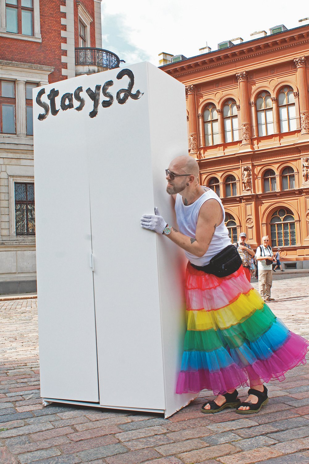 aikinas su vaivorykštiniu sijonu stumia baltą spintą užrašytą „Stasys2“.