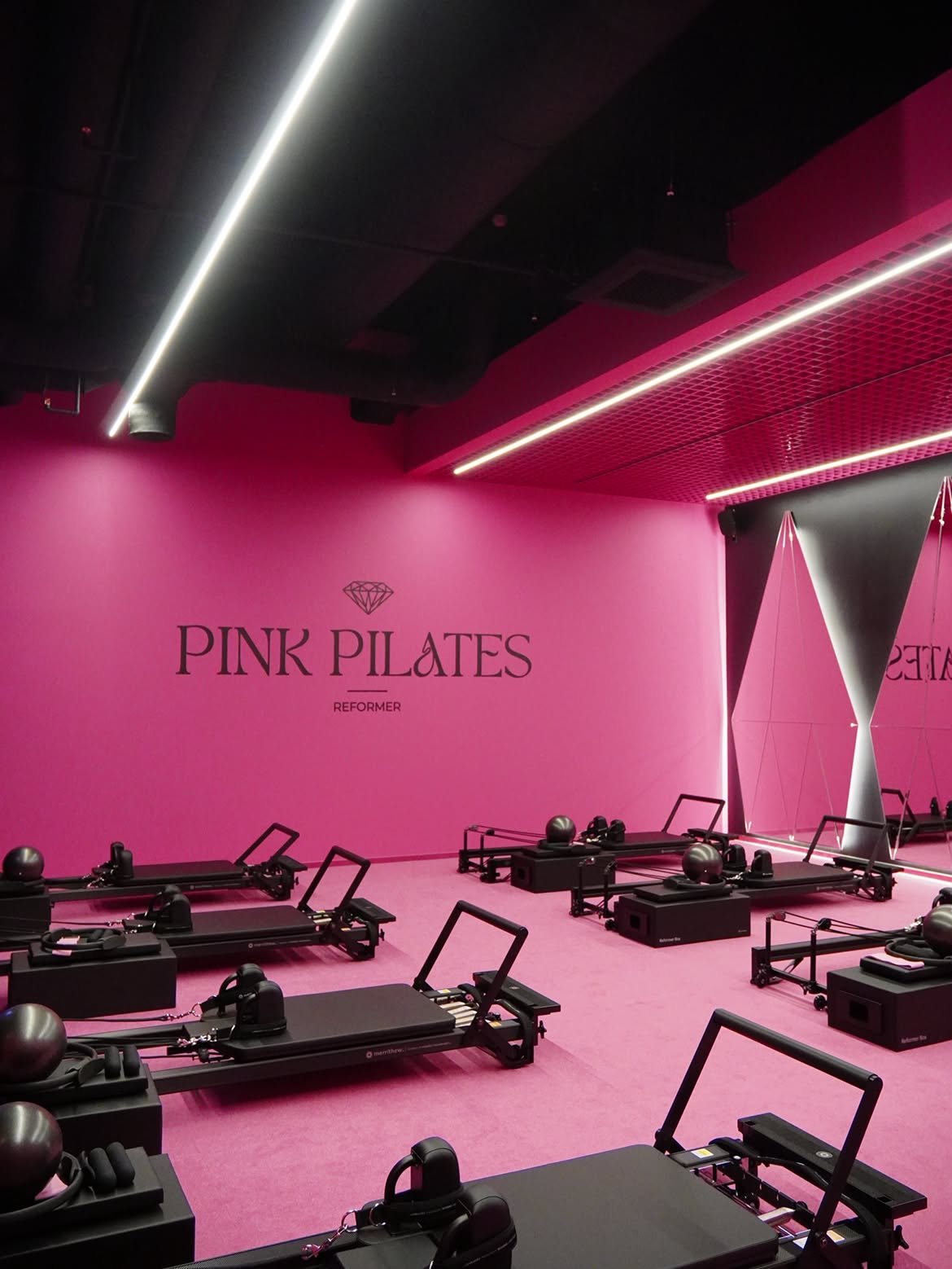 Rožinė pilateso studijos salė su reformerstakais ir „Pink Pilates“ užrašu