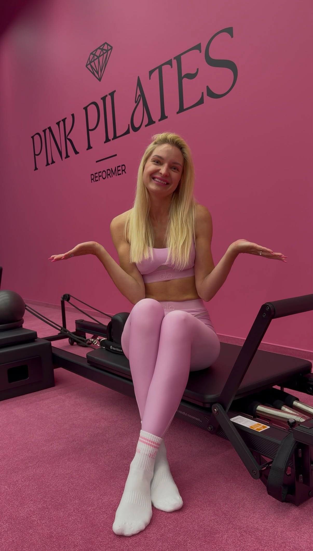 Šviesiaplaukė moteris su rožine sportine apranga sėdi ant pilateso reformerio