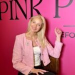 Besišypsanti šviesiaplaukė moteris su rožiniu švarkeliu prie „Pink Pilates“ sienelės