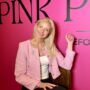 Besišypsanti šviesiaplaukė moteris su rožiniu švarkeliu prie „Pink Pilates“ sienelės